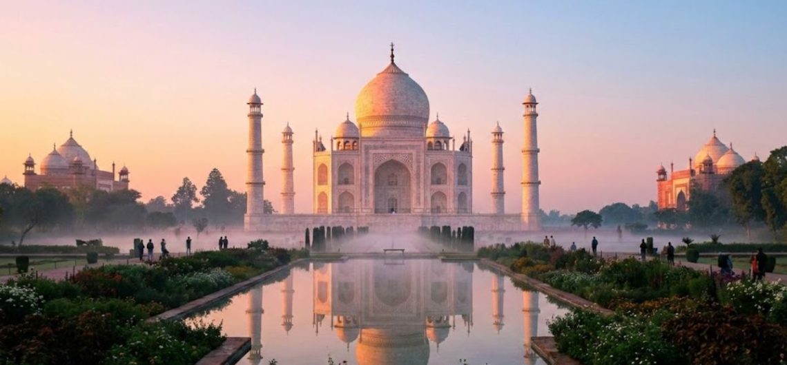 A wide, scenic view of the white marble Taj Mahal at sunrise, glowing with golden light and perfectly reflected in the water of a long pool in the foreground under a pastel-colored sky.